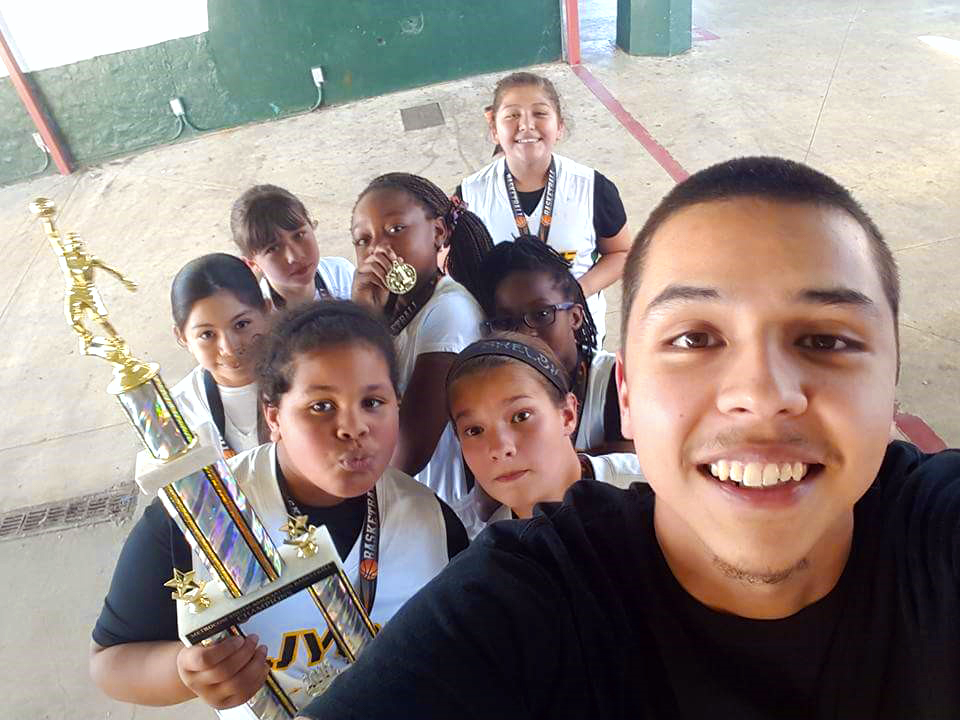 A Terry Scholar is smiling for a selfie with a group of kids, one holding a large trophy, in the background