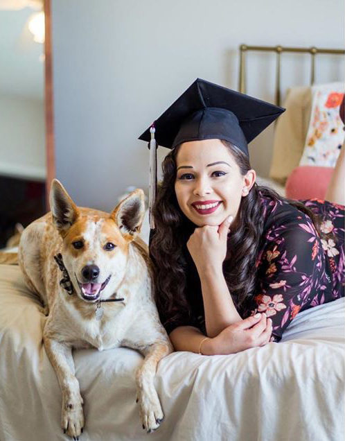 A TWU Terry Scholar laying on a bed next to a medium-sized white and tan dog. Her hand is posed under her chin,as she smiles wearing a black graduation cap