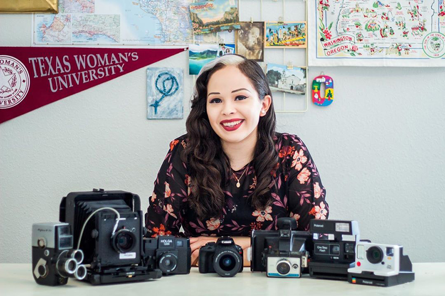 Terry Scholar in a floral print long sleeved dress is seated, smiling, at a desk with many different types of cameras displayed in front. She has long black hair with a single white streak of hair in front. To the left in the background is a pennant that reads Texas Women's University